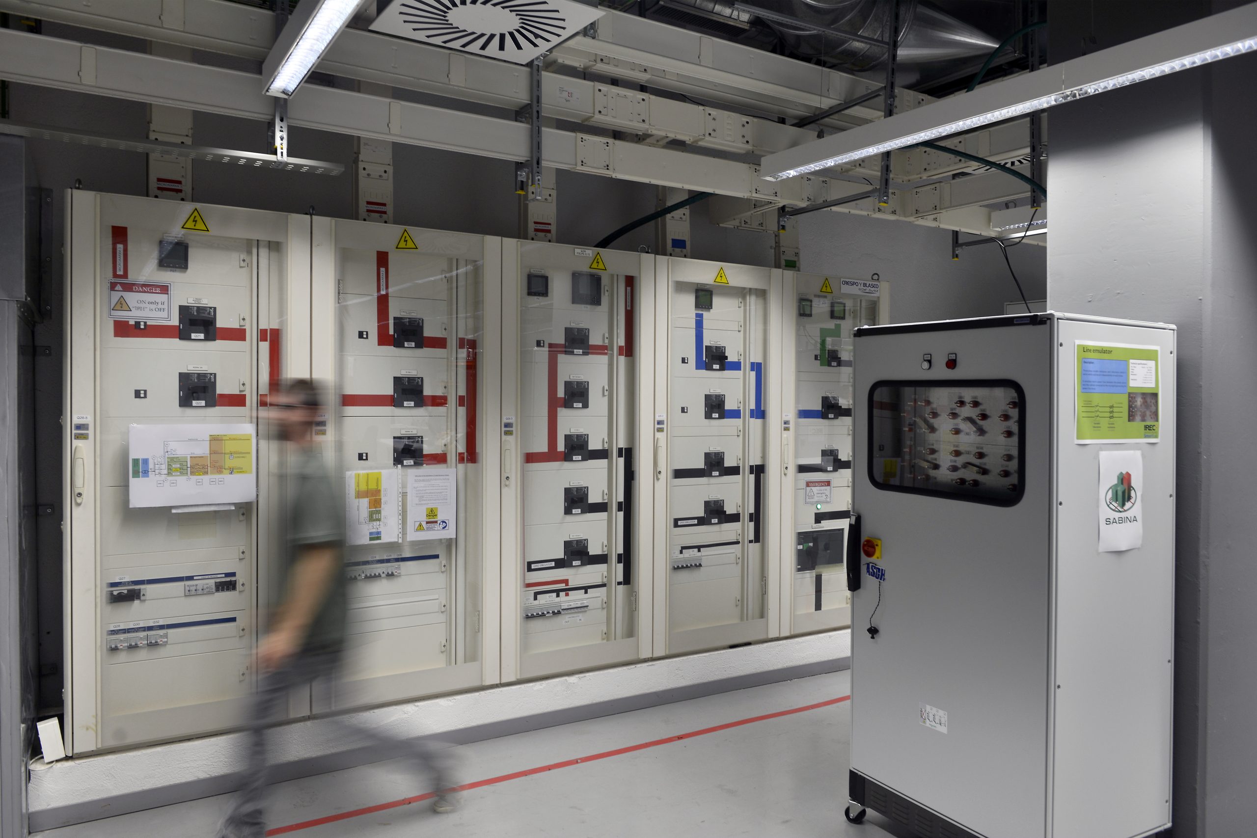 Person walking in front of industrial laboratory machinery, representing the IREC SmartLab experimental infrastructure for testing integrated energy systems, microgrids, batteries and energy management technologies.