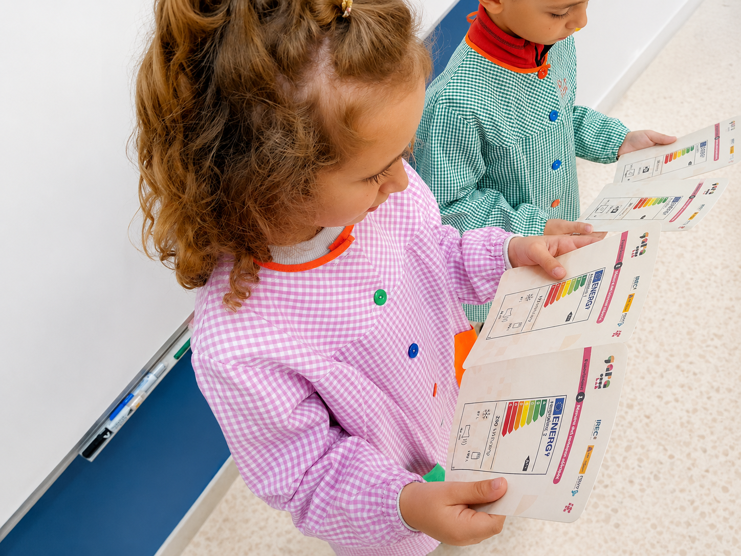 High-angle view of two schoolchildren in school smocks handling and reviewing educational cards with energy efficiency graphs as part of the learning activities from the pedagogical suitcases.