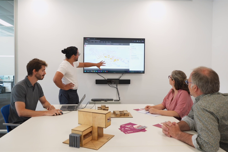 Four people view an urban visualisation on a wall-mounted screen, with architectural models on the table.